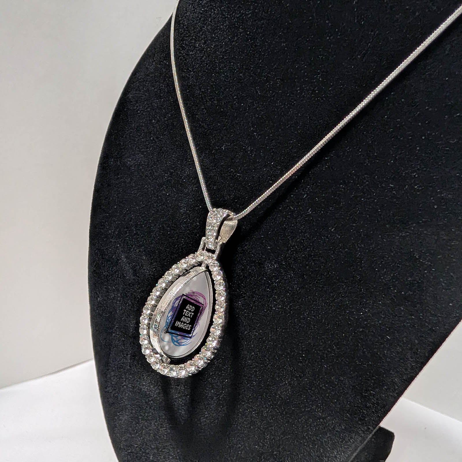 Customizable double-sided teardrop shaped silver rhinestone pendant with a silver necklace on a black stand